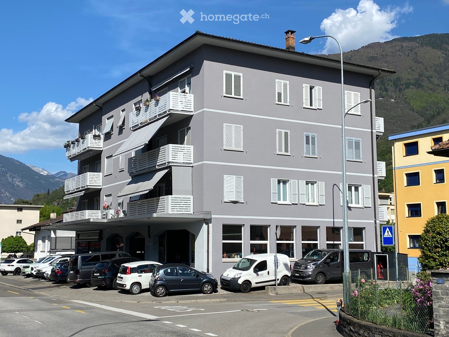 Building. A multi-story residential building with grey exterior, white-framed windows, and balconies, located in a town with mountains in the background.