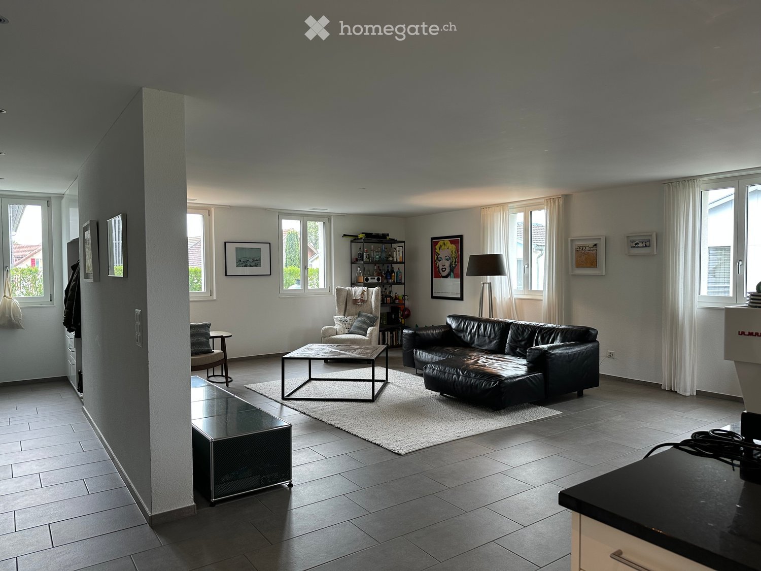 Living Room. Modern living room with large windows, grey leather sofa, coffee table, armchair, side table, floor lamp, and shelves with decorative items.