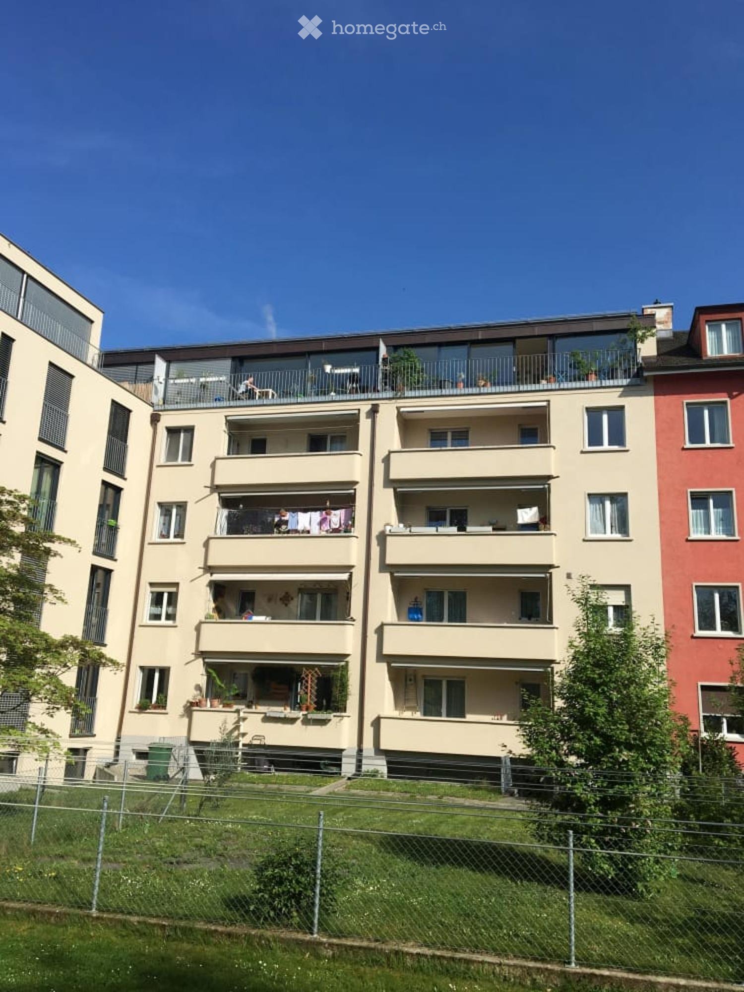 Apartment building, beige facade, several balconies, greenery, chain link fence, trash bins, red building on the right