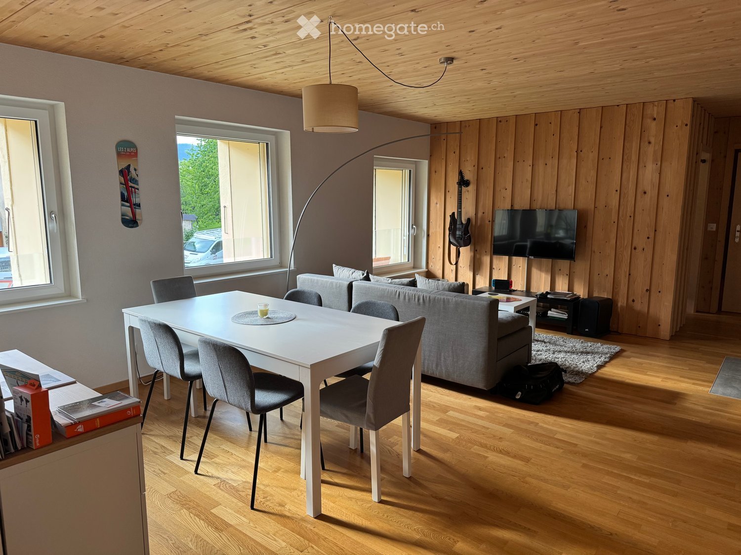 Modern living room with wooden floors, large windows, a grey sofa, a white dining table with chairs, a hanging lamp, a floor lamp, a guitar on the wall, and a television mounted on the wall.