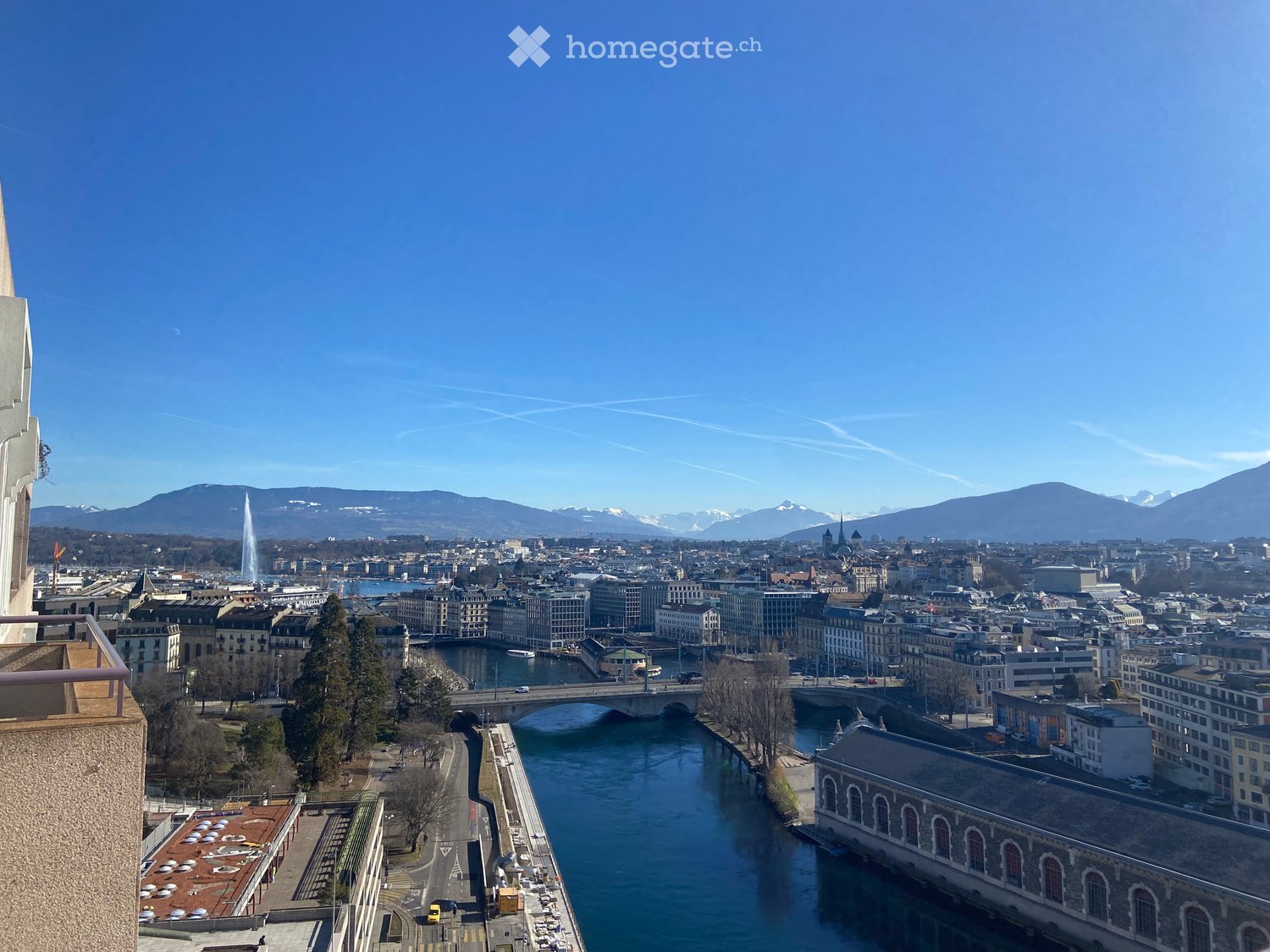 Panoramic cityscape view from a balcony, featuring a river, bridges, buildings, and distant mountains.