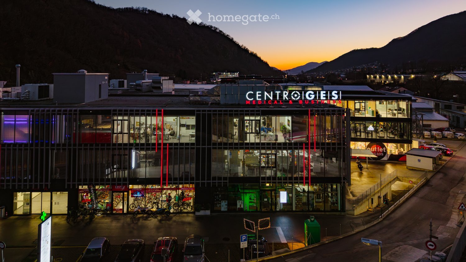 Modern multi-story commercial building with large glass windows, illuminated signage reading "CENTRO GIESI MEDICAL & BUSINESS," and visible interior spaces including offices and retail areas. The building is surrounded by parked cars and bicycles, with a