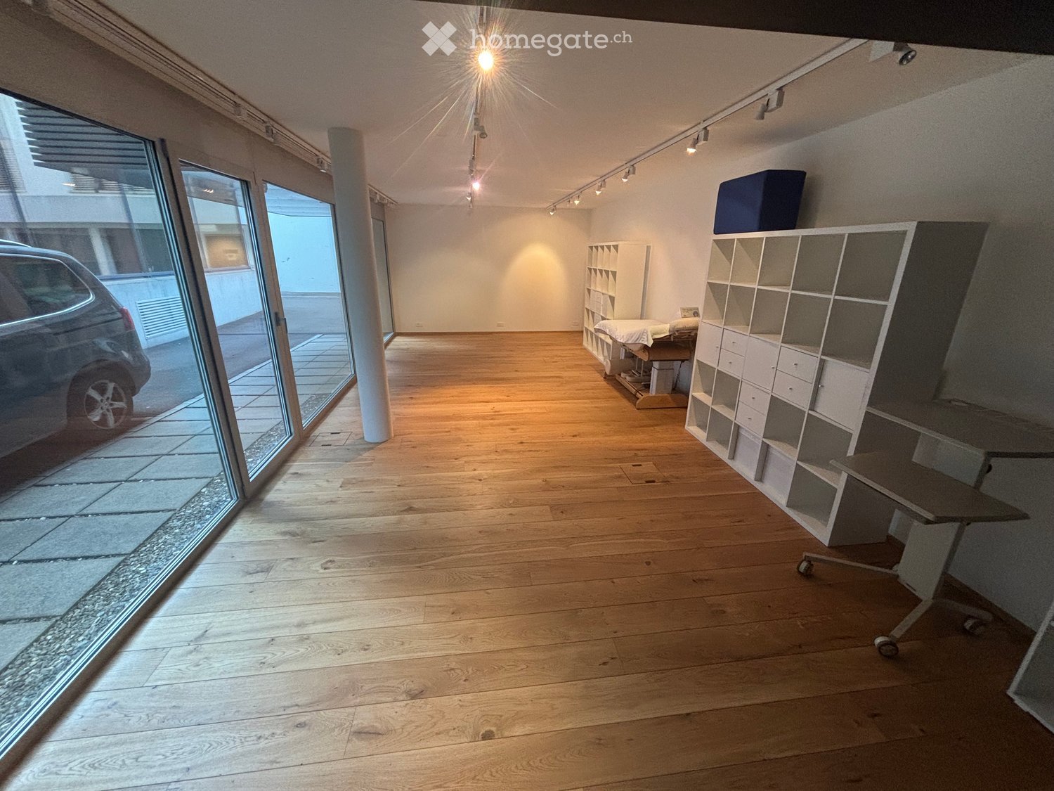 Bright, open room with wooden flooring, large glass sliding doors leading outside, white shelving units, a small table, and a stool.