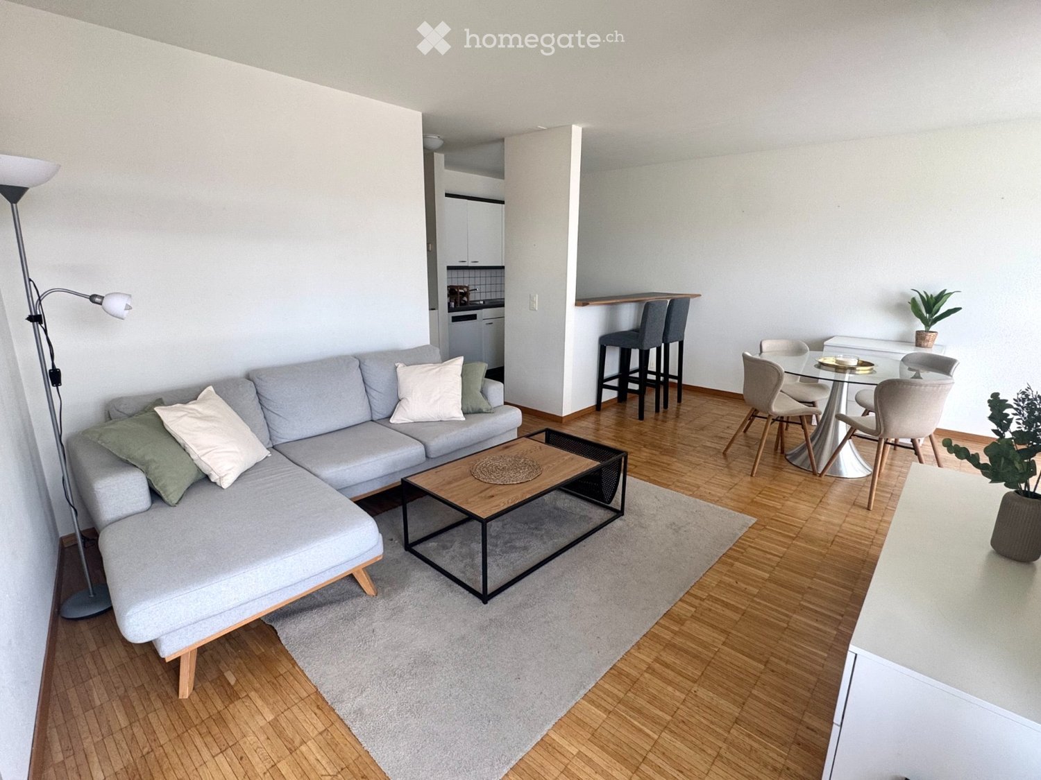 Modern living room with grey sectional sofa, wooden coffee table, dining area with glass table and chairs, and bar stools near the kitchen.