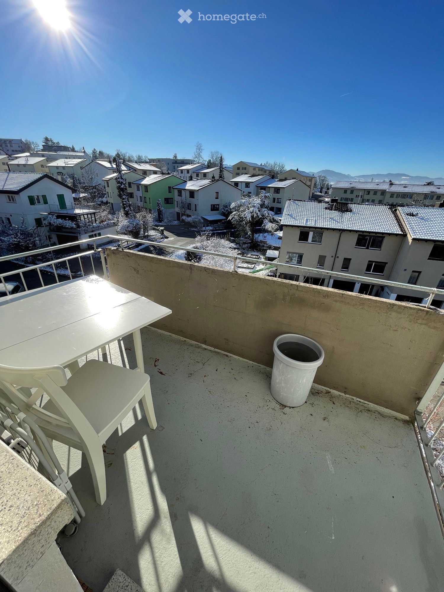 Balcony / Terrace. Balcony with a white table, two white chairs, and a white trash can, overlooking a residential area with snow-covered trees and houses.