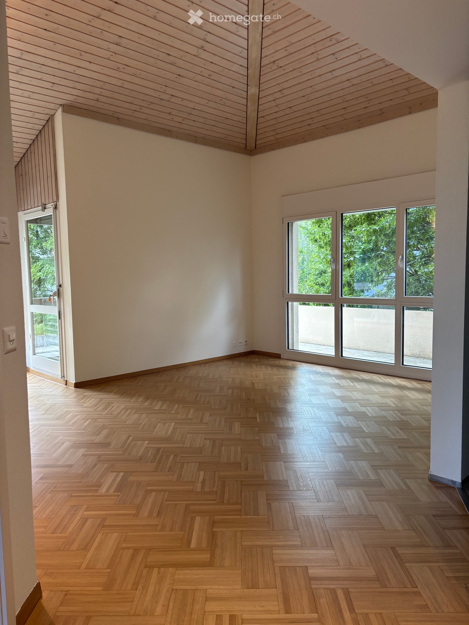 Empty room with wooden herringbone flooring, white walls, large windows, and a sliding glass door leading outside.
