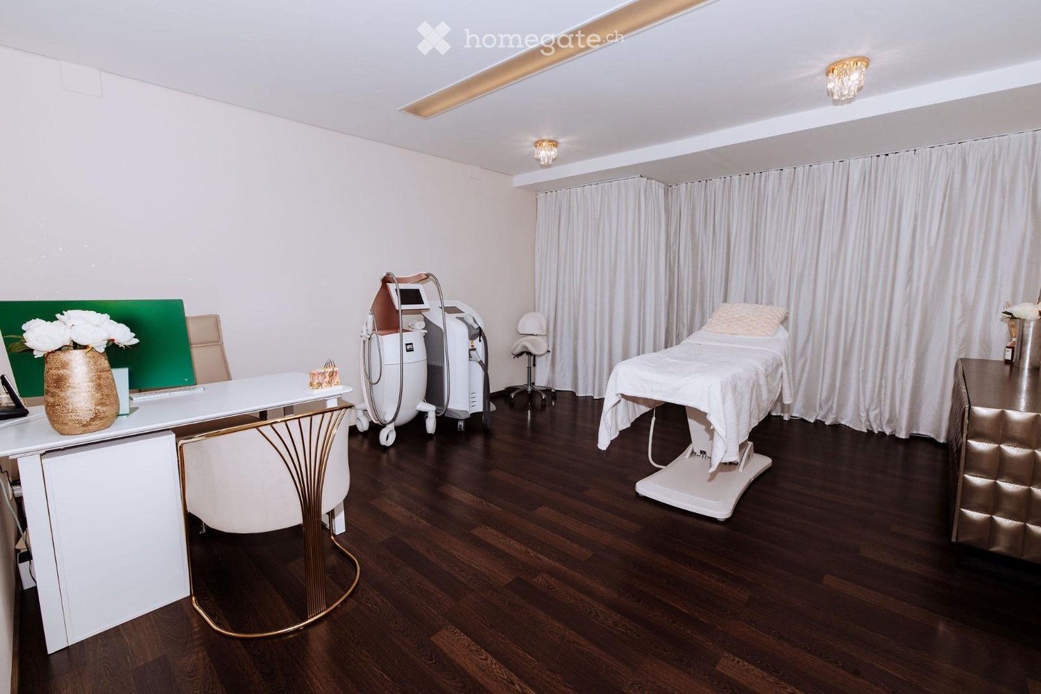 Modern, well-lit room with dark wooden flooring, white walls, and a treatment table with a white sheet. A desk with a vase of white flowers, a chair, and a medical device are visible.