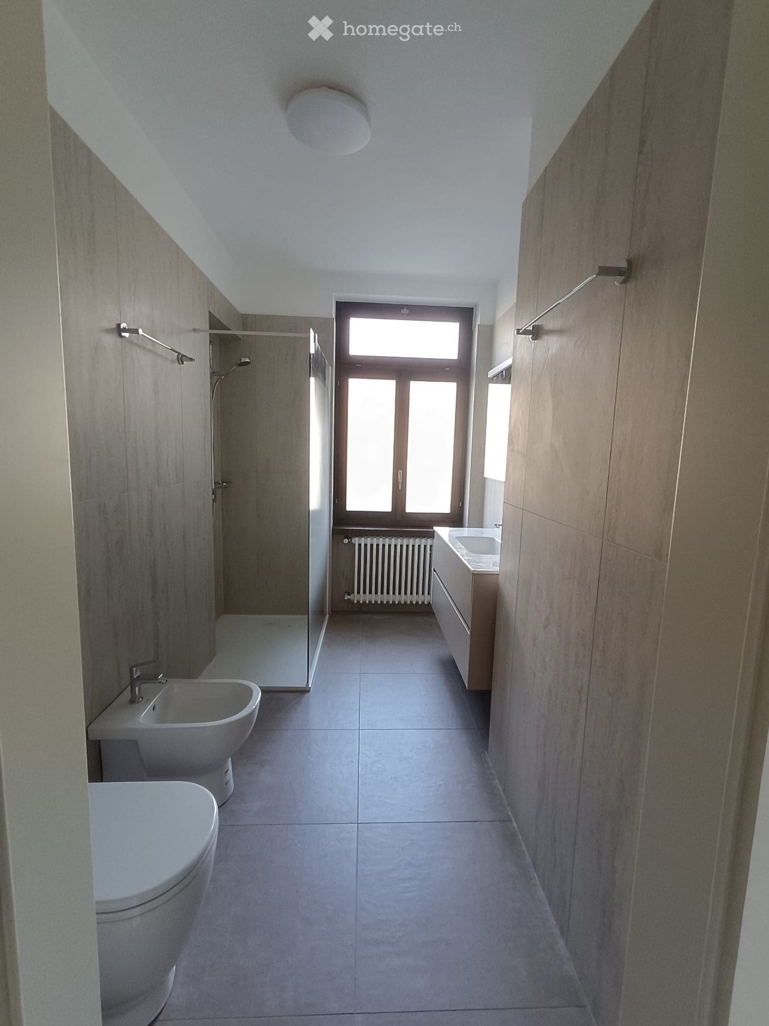 Modern bathroom with a toilet, sink, shower, and radiator. Features light-colored tiles and a large window providing natural light.