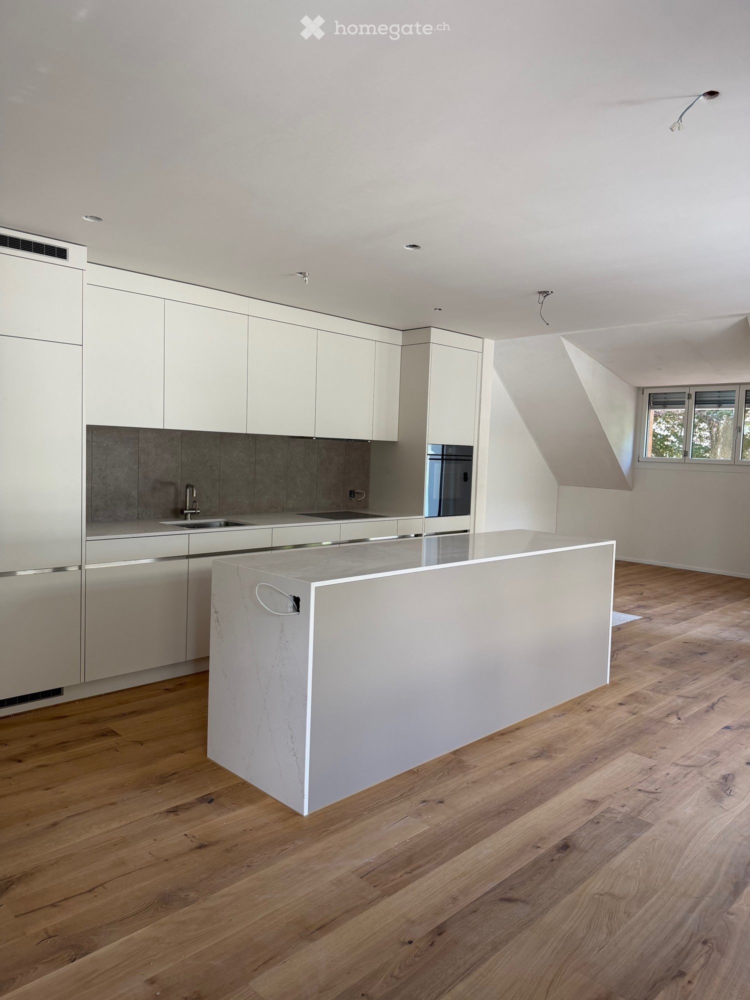 Modern kitchen with white cabinets, a central island, and a sink area. The floor is made of light wooden planks, and the backsplash is a dark grey material.