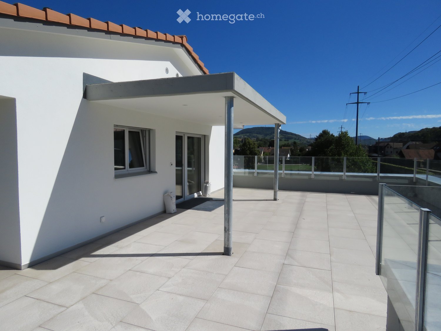 Modern balcony with light-colored tiles, glass railing, and a view of the surrounding landscape.