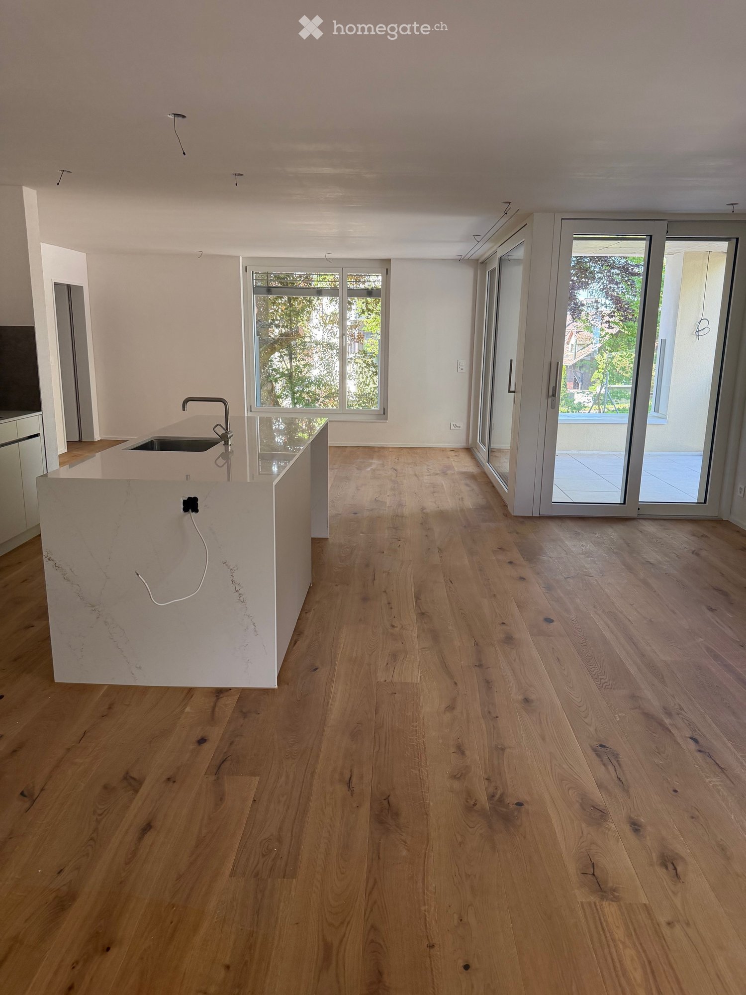 Empty room with light wooden flooring, white walls, and a modern kitchen island with a sink. Large windows and sliding glass doors lead to an outdoor area.