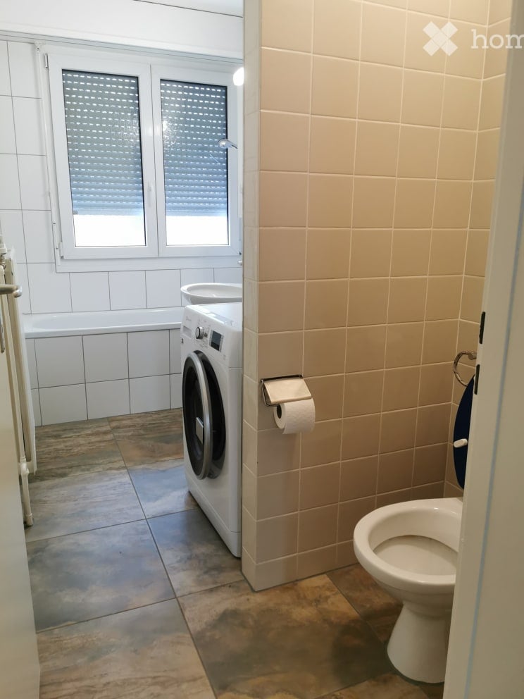 Bathroom with a toilet, washing machine, bathtub, and toilet paper holder. The walls are tiled, and there is a window with blinds.