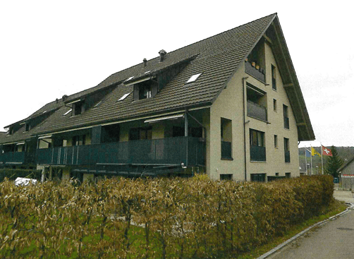 A multi-story residential building with a sloped roof, multiple windows, and balconies. The building is surrounded by a hedge and a paved driveway.