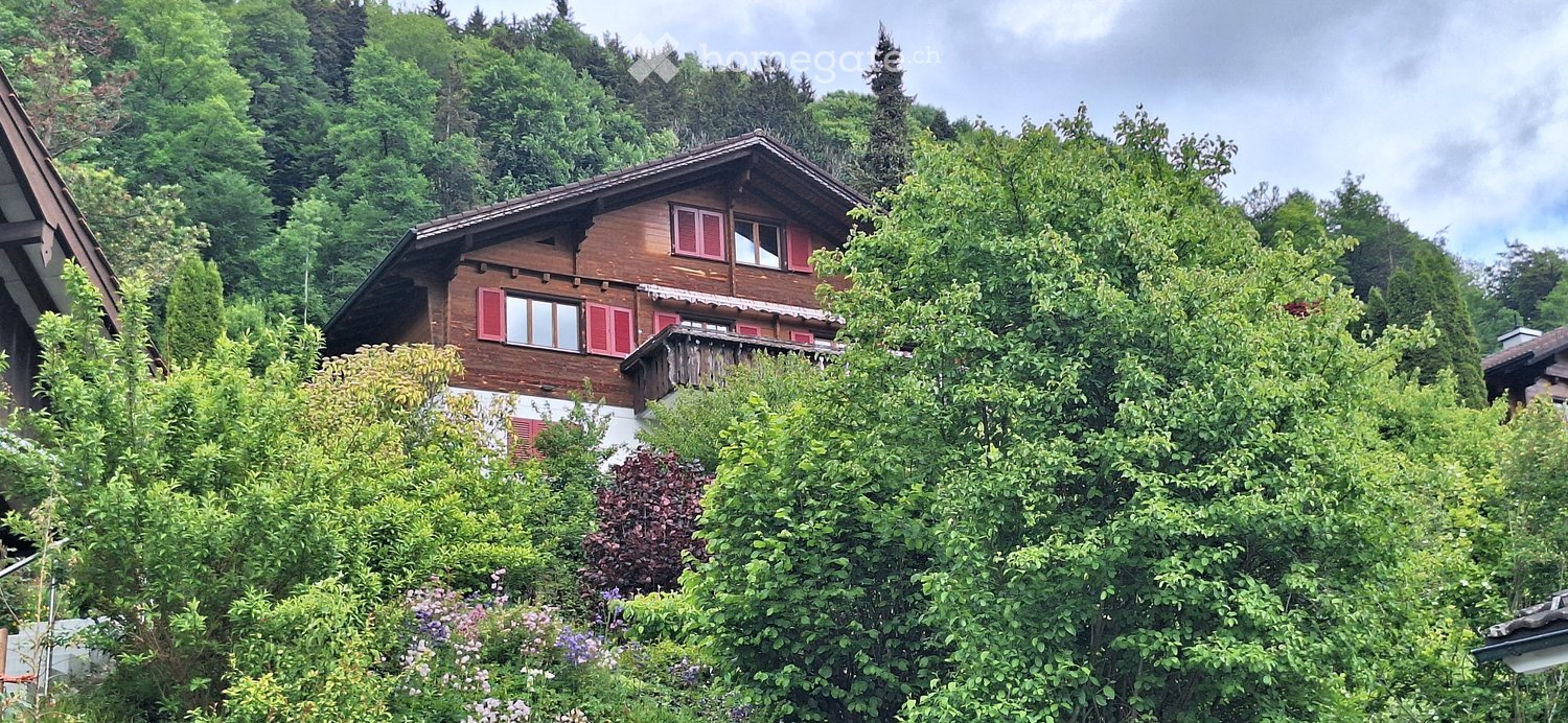 Traditional wooden house with red shutters and a balcony, surrounded by lush greenery and trees.