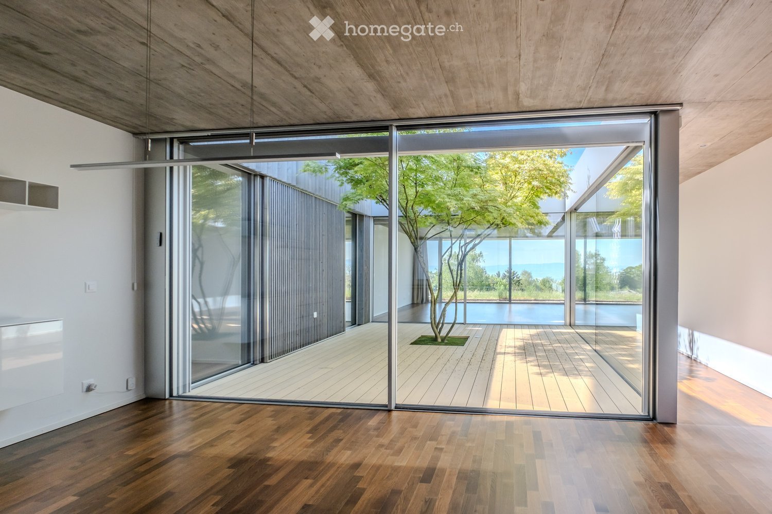 Modern hallway with wooden flooring, large glass sliding doors leading to an outdoor deck, visible tree, and swimming pool in the background.