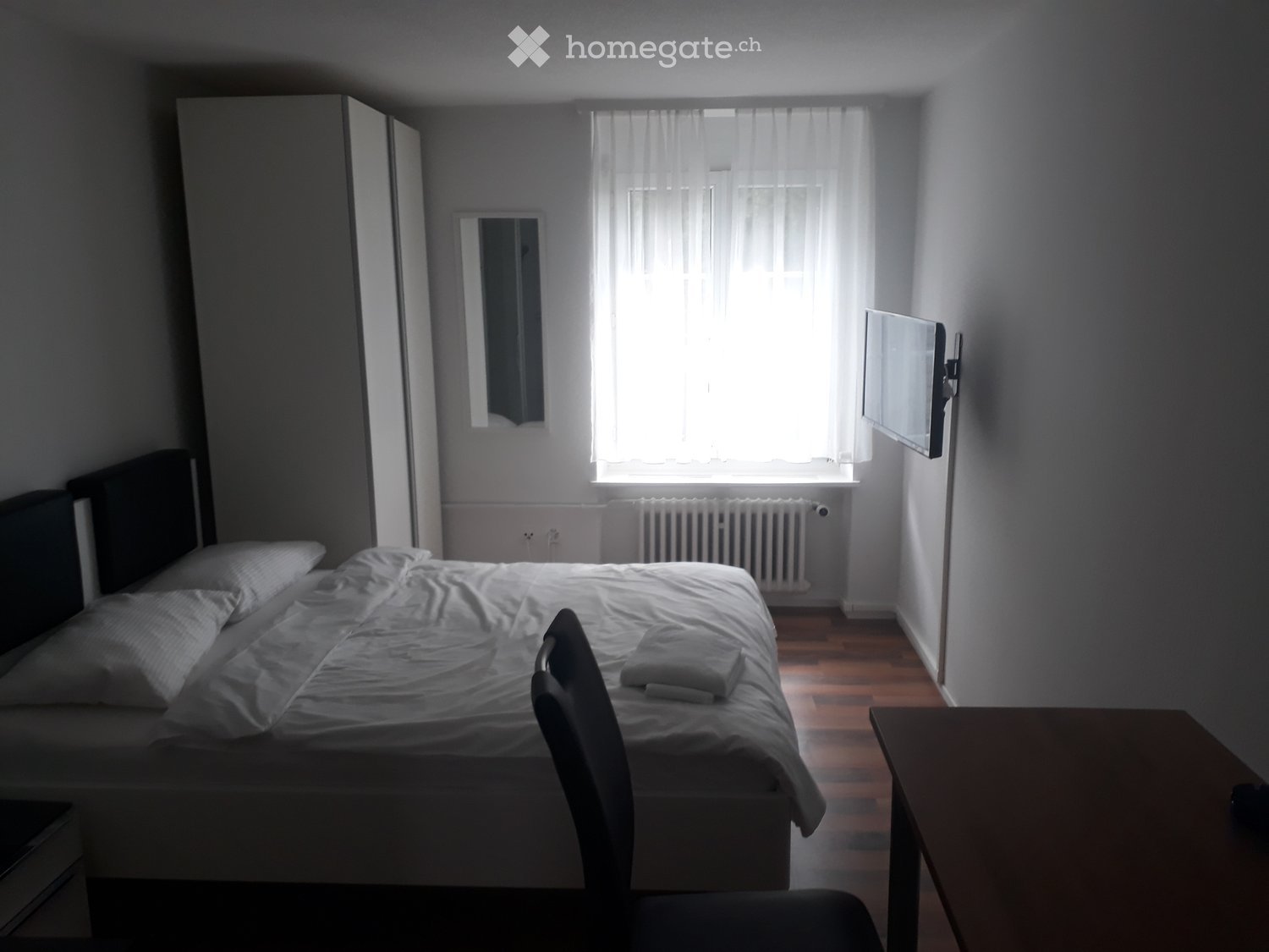 A well-lit bedroom with a double bed, a chair, a small table, a mirror, a wardrobe, and a wall-mounted TV. The room has wooden flooring and large windows with sheer curtains.