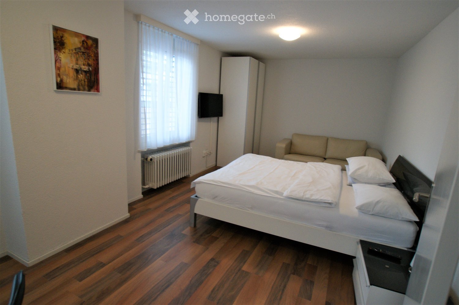 A compact bedroom with a double bed, a sofa, a television mounted on the wall, a wardrobe, and a picture frame on the wall. The room features wooden flooring and large windows with white curtains.