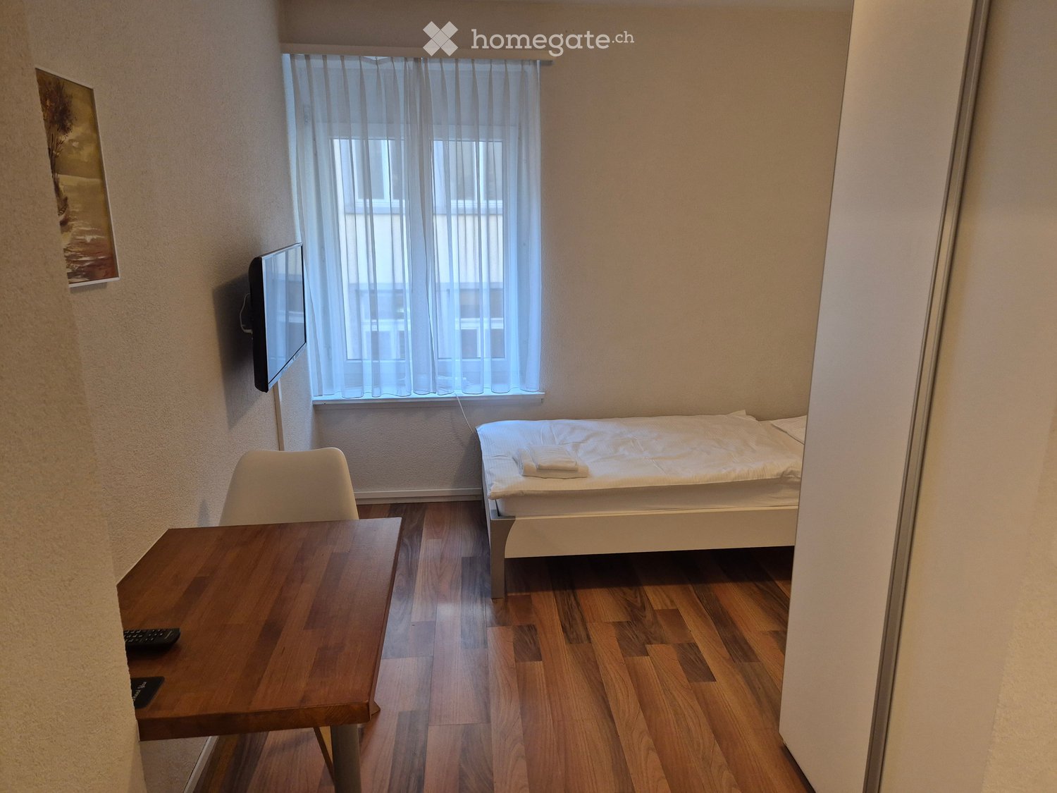 Cozy bedroom with a single bed, wooden table, chair, and wall-mounted TV. Light-colored walls and wooden flooring.