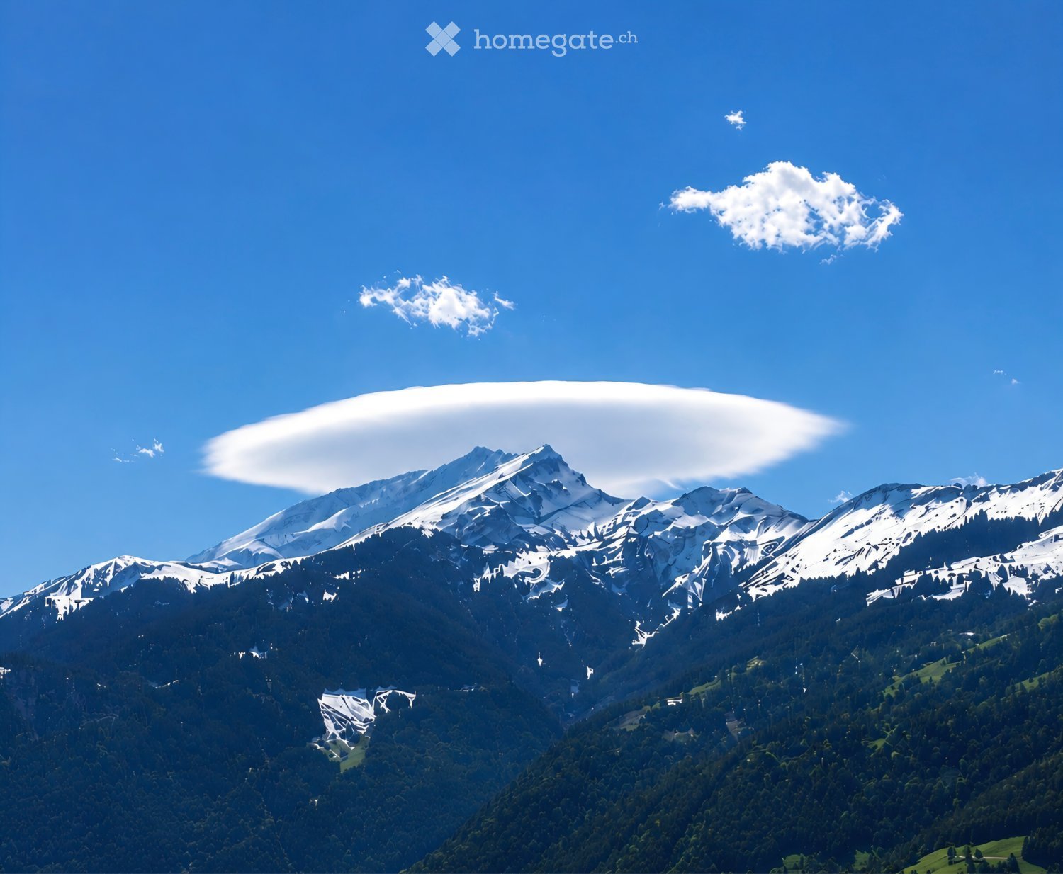 A panoramic view of snow-capped mountain peaks under a clear blue sky with scattered clouds, surrounded by dense forest and green valleys below.