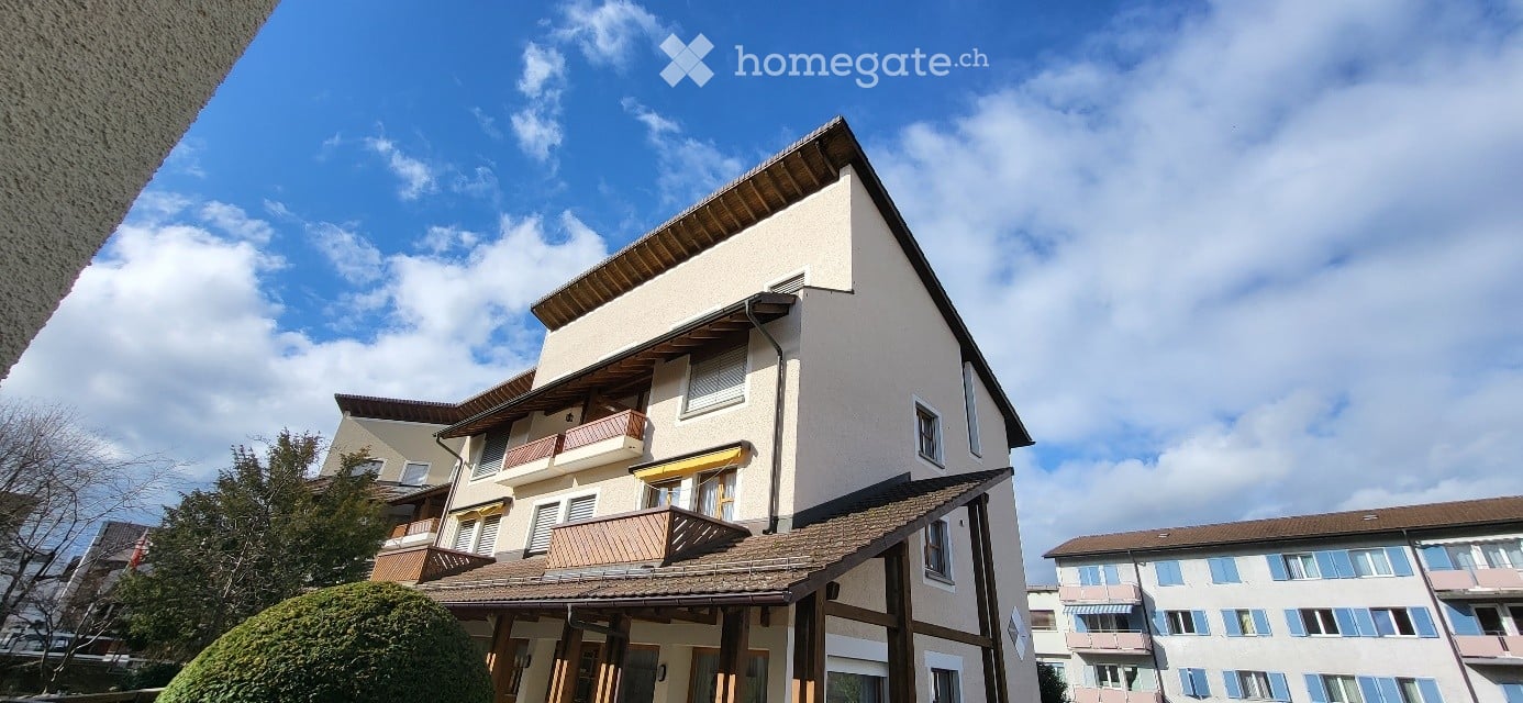 Two-story residential building with balconies, wooden shutters, and a tiled roof under a partly cloudy sky.