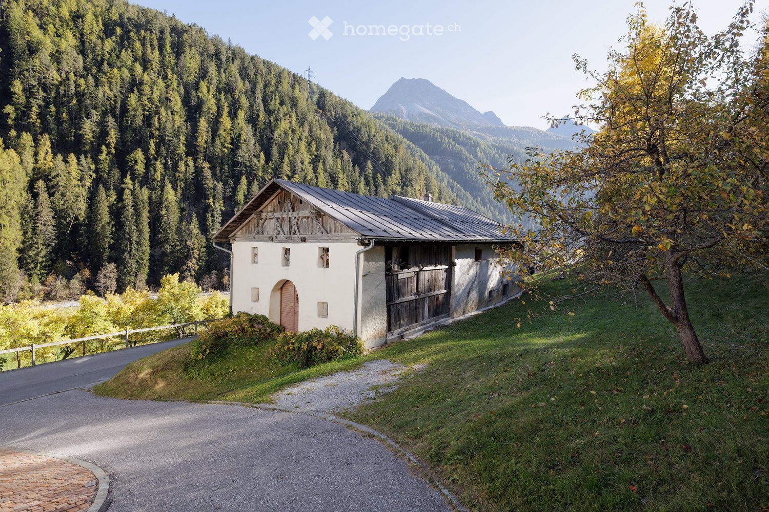 A small, rustic wooden house with a stone base, situated in a lush green valley surrounded by dense forests and mountains.