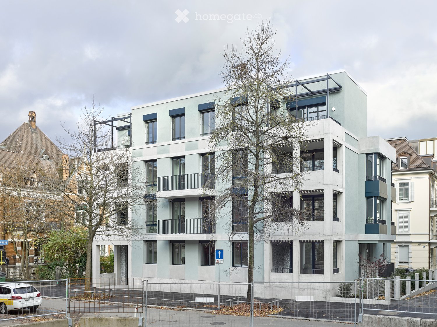 Modern multistory building with light gray exterior, multiple windows, balconies, fenced-in entrance area