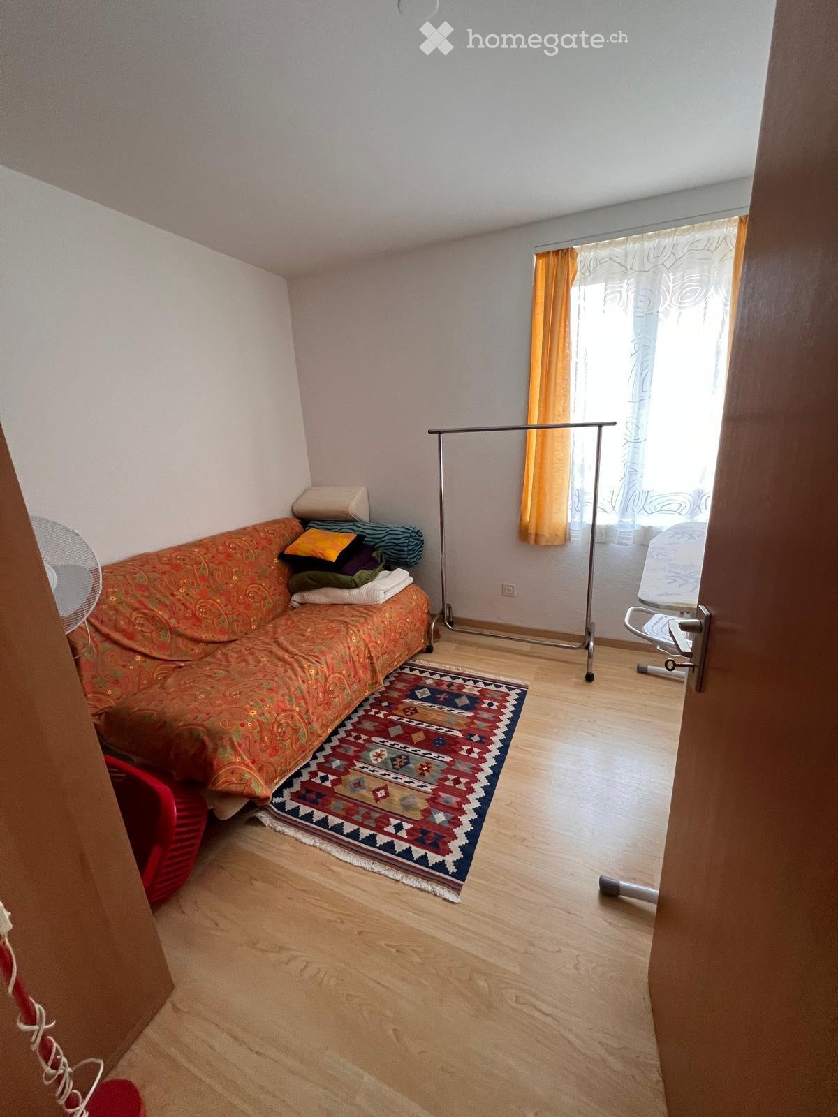 A small, cozy living room with a patterned sofa, a colorful rug, a fan, a curtain, and a clothes rack.