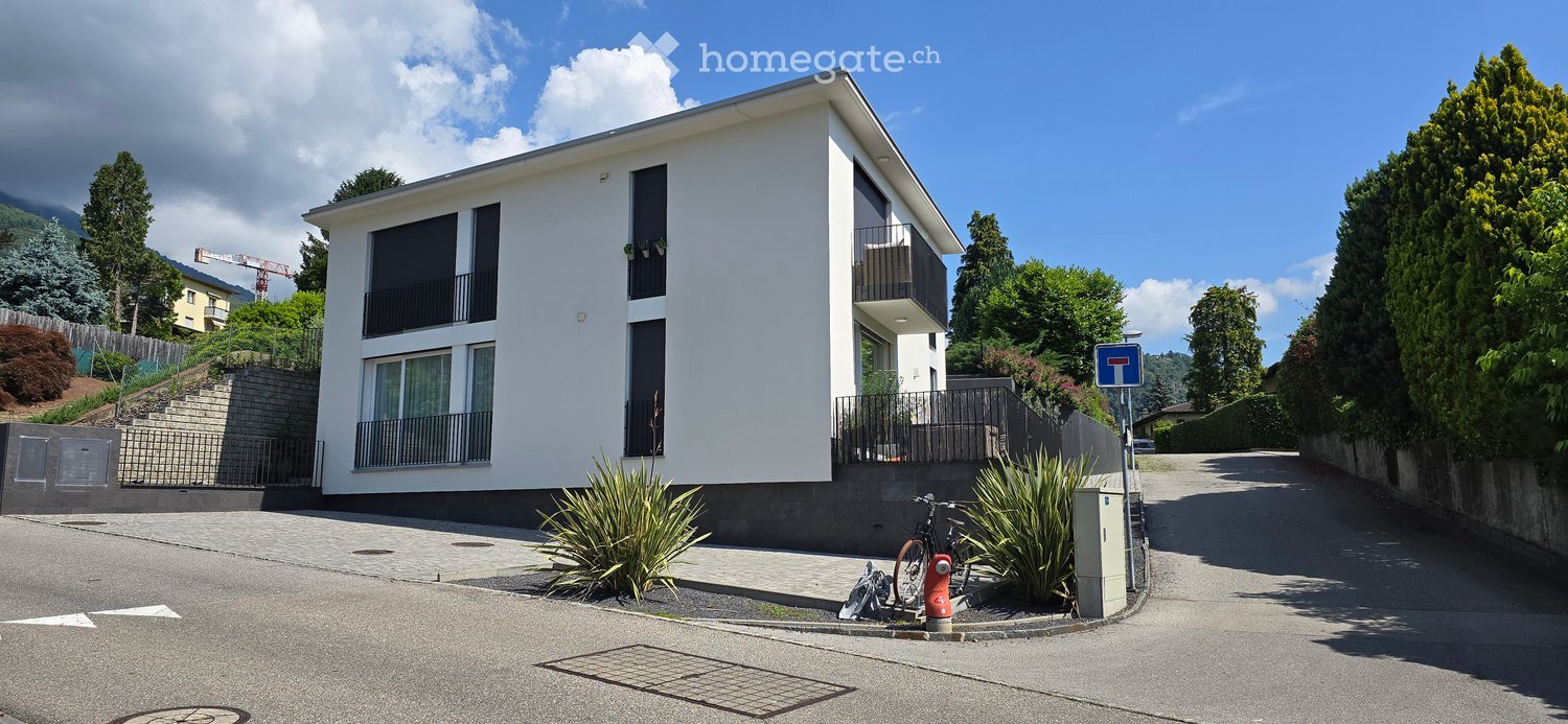 Modern two-story house with white exterior, large windows, and balconies. The house is situated on a raised ground floor with a paved driveway and surrounding greenery.