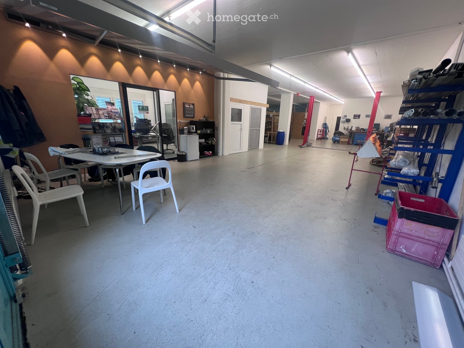 A spacious, open-plan room with a concrete floor, large windows, and various furniture including tables, chairs, shelves, and office equipment.