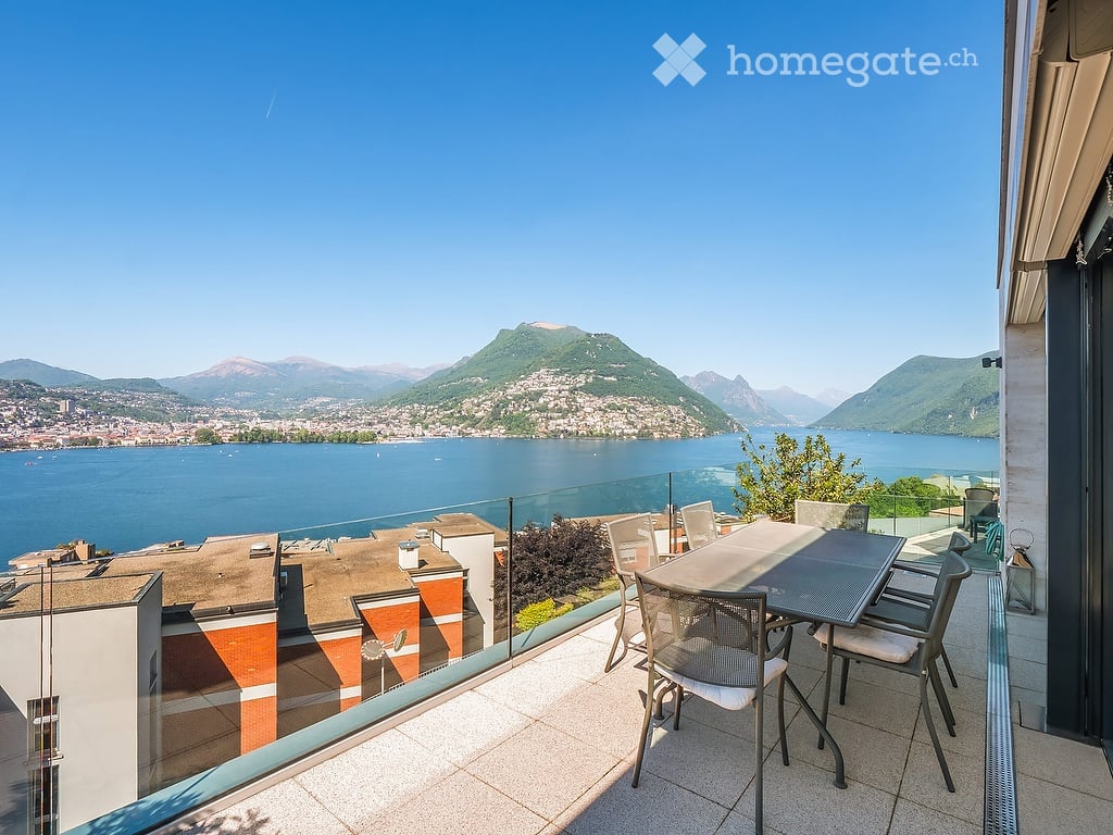 Balcony with a scenic view of a lake and mountains, featuring a table and chairs.