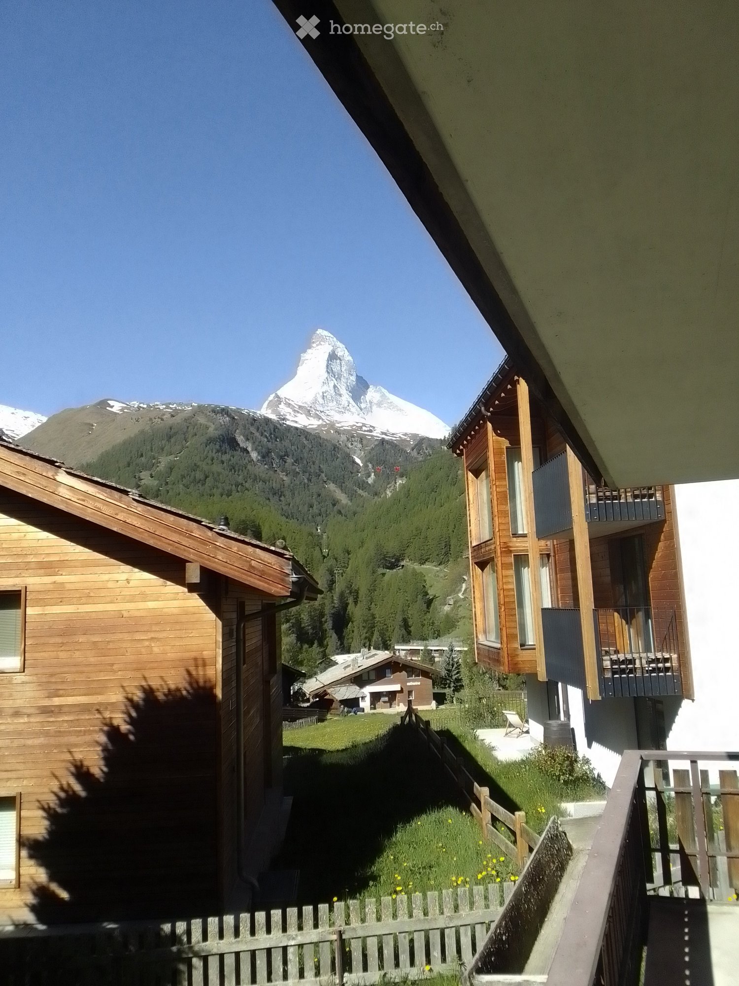 View of a picturesque alpine village with wooden houses, a grassy area, and a stunning mountain backdrop featuring the Matterhorn.