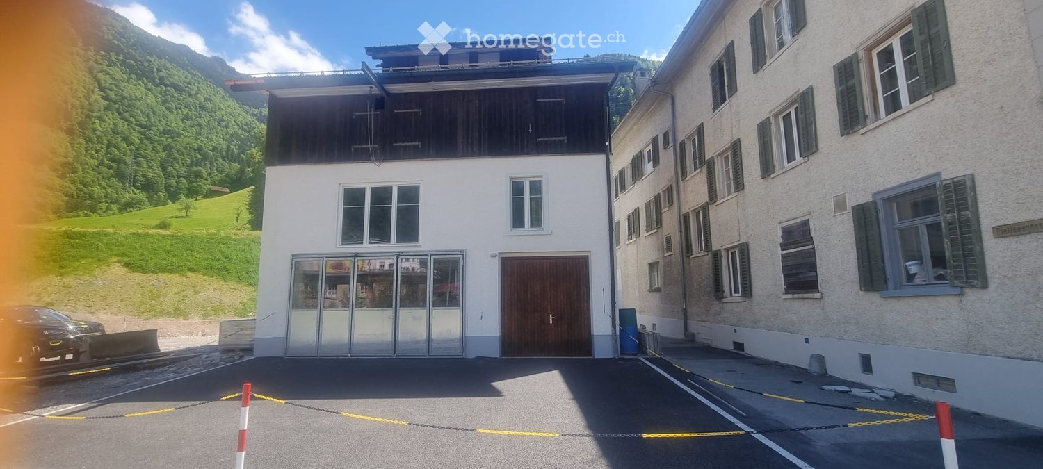 Two-story building with white facade, large windows, and a wooden garage door. Adjacent building has multiple windows with shutters. The scene includes a parking area with a car and a scenic mountain backdrop.