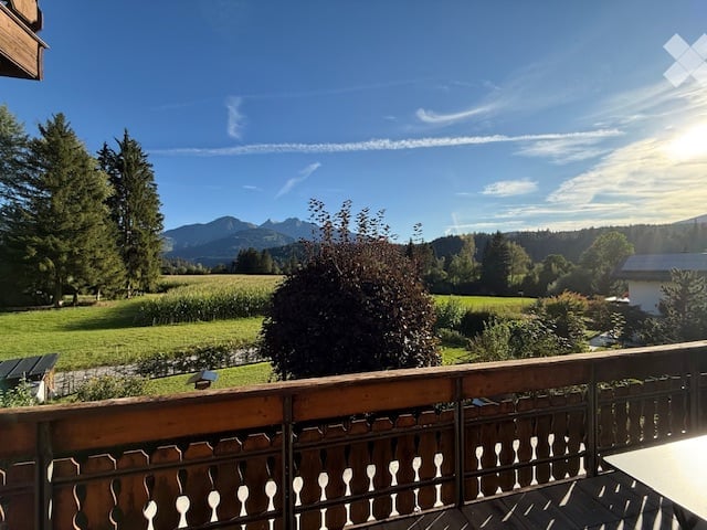 Wooden balcony with railings, wooden table and chairs, scenic view of a field, trees, and mountains