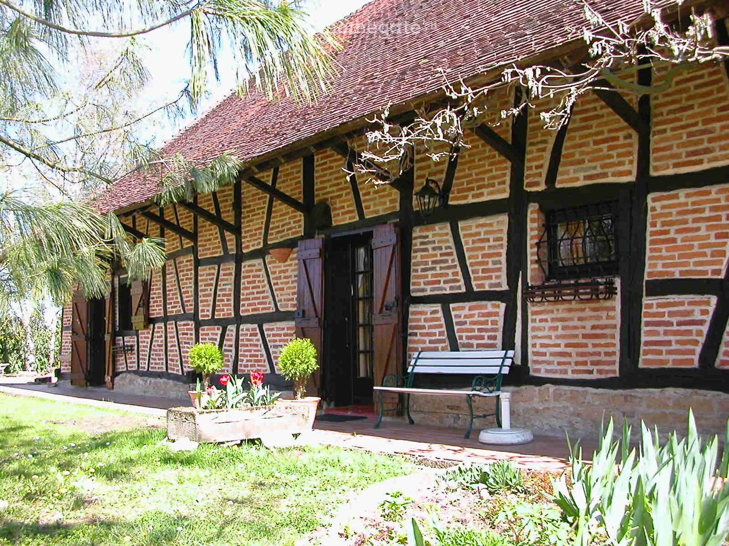 Two-story brick house with a tiled roof, black timber framing, and a covered porch area with a bench and potted plants.