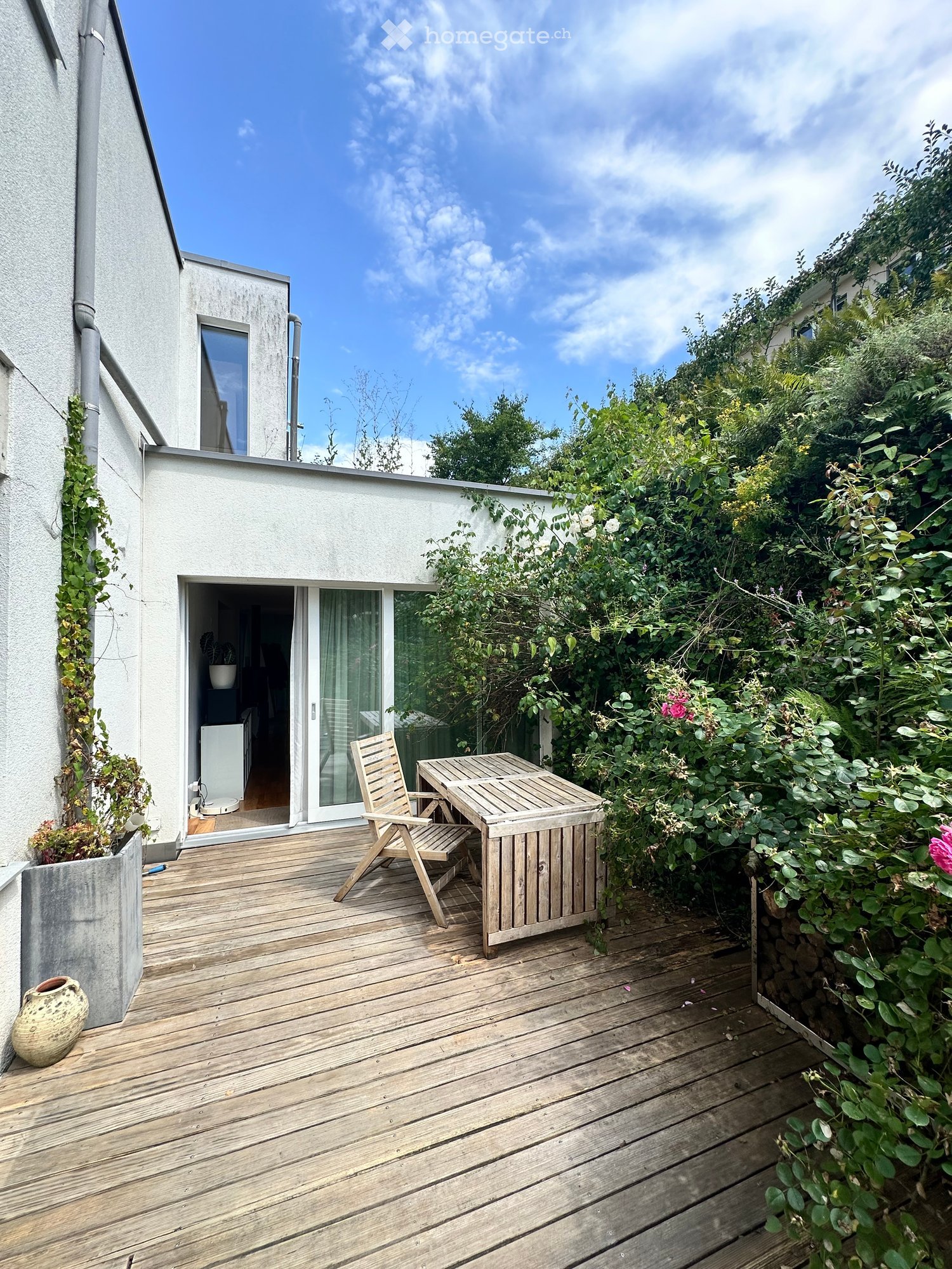 Wooden deck with a table and chair, surrounded by greenery and a view of the building.