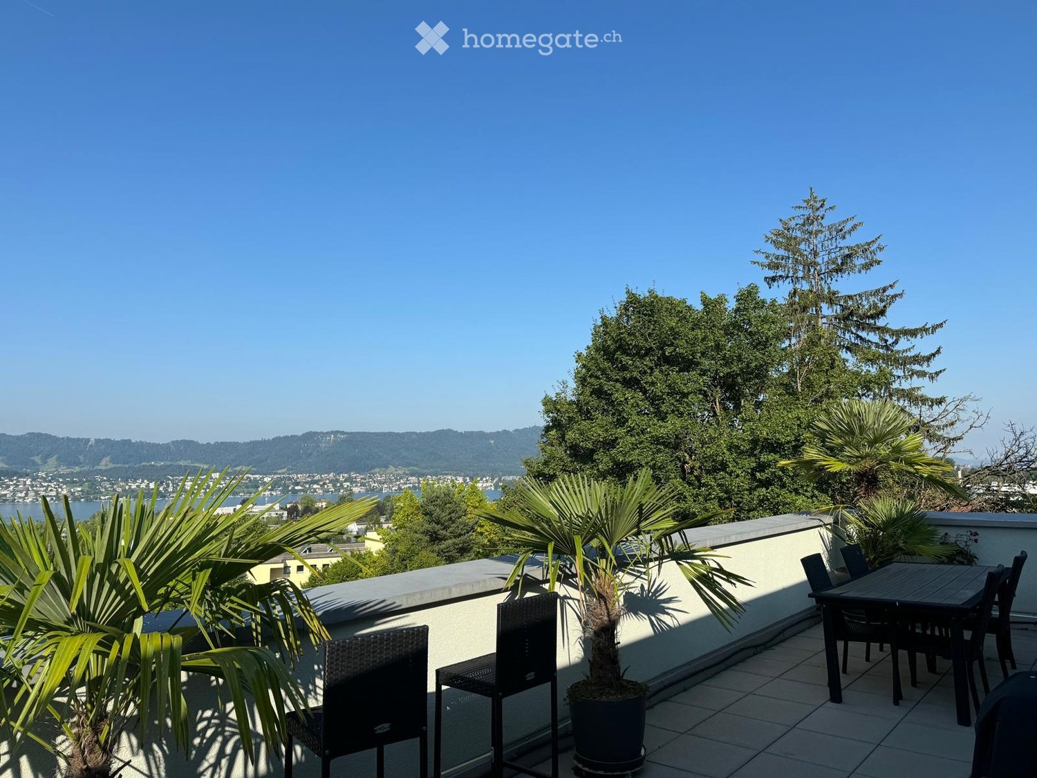 Balcony with tiled floor, potted plants, and outdoor furniture including a table and chairs, overlooking a scenic view of a city and water.