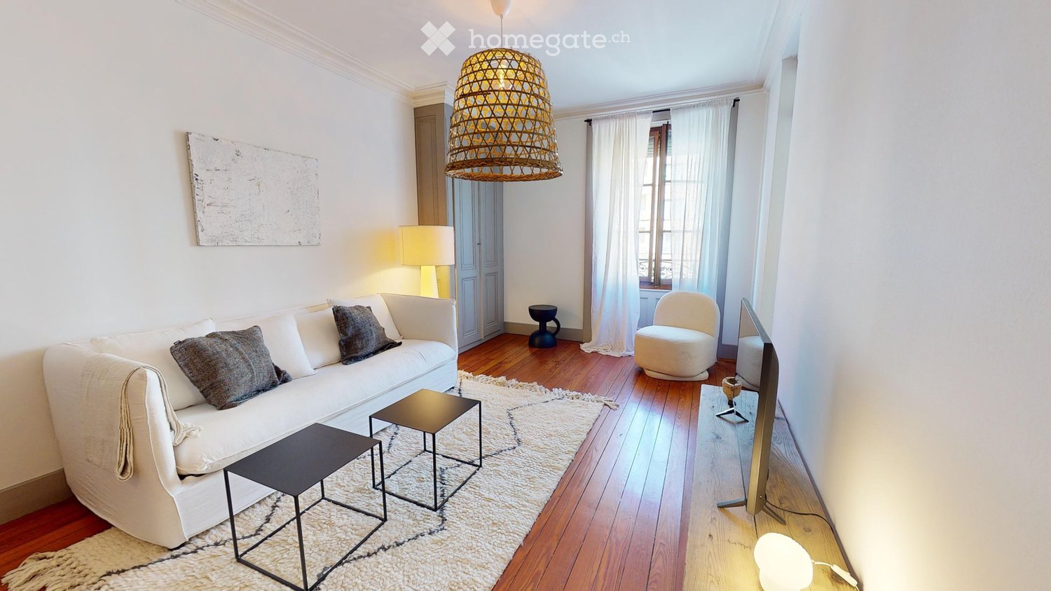 Modern living room with white walls, wooden flooring, a white sofa with pillows, a small black table, a white armchair, a hanging woven lamp, and a television on a stand.