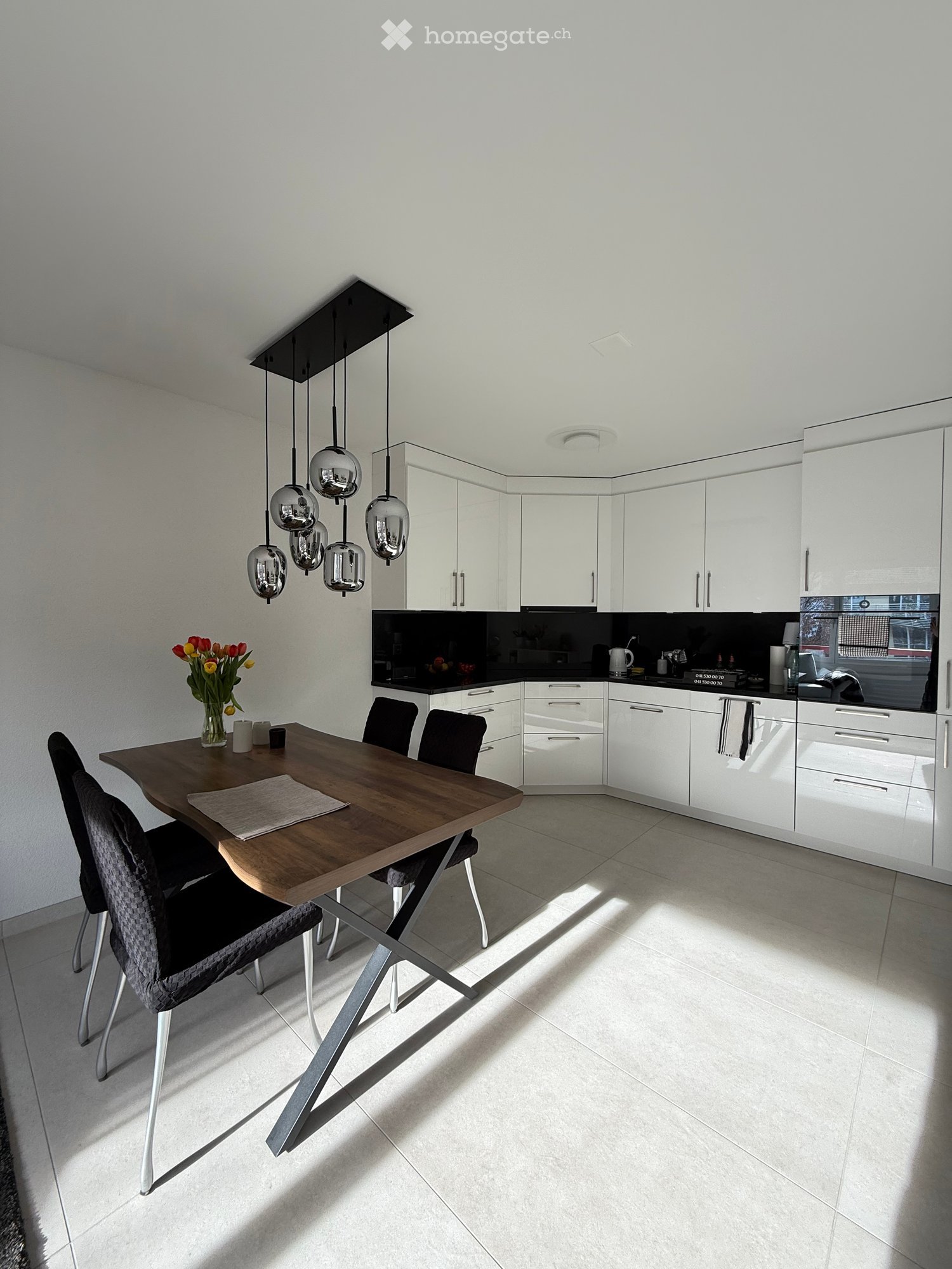 Modern kitchen with white cabinets, a wooden dining table, pendant lights, and a vase with flowers on the table.