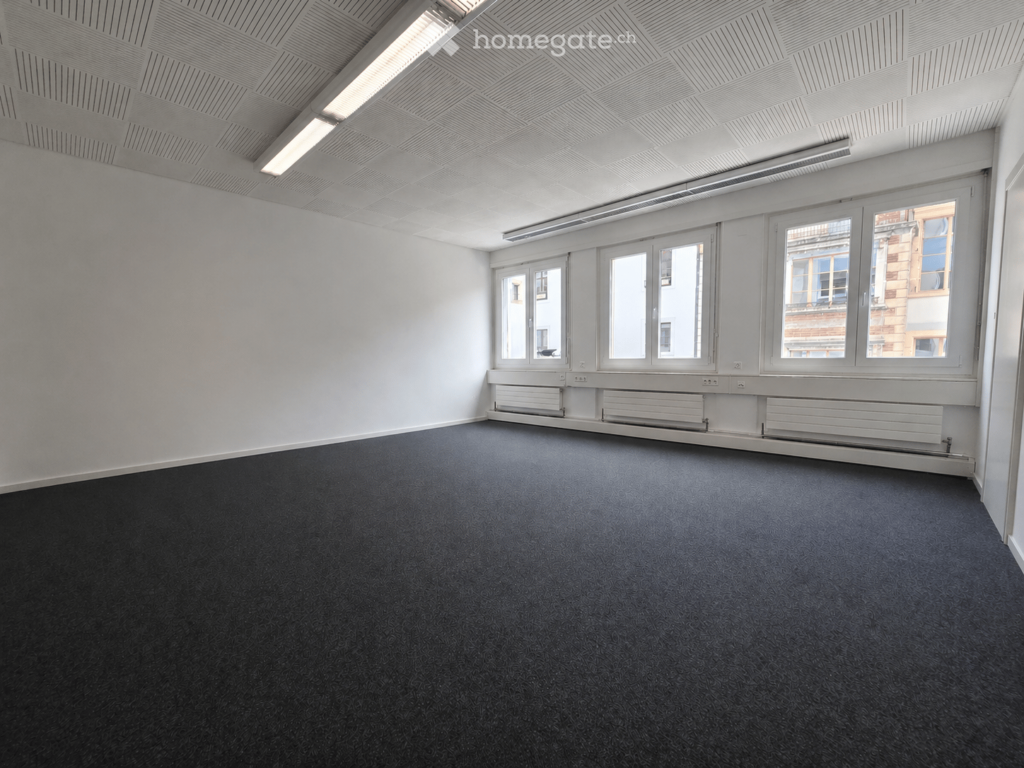 Empty room with white walls, dark carpeted floor, large windows, and white ceiling with recessed lighting.