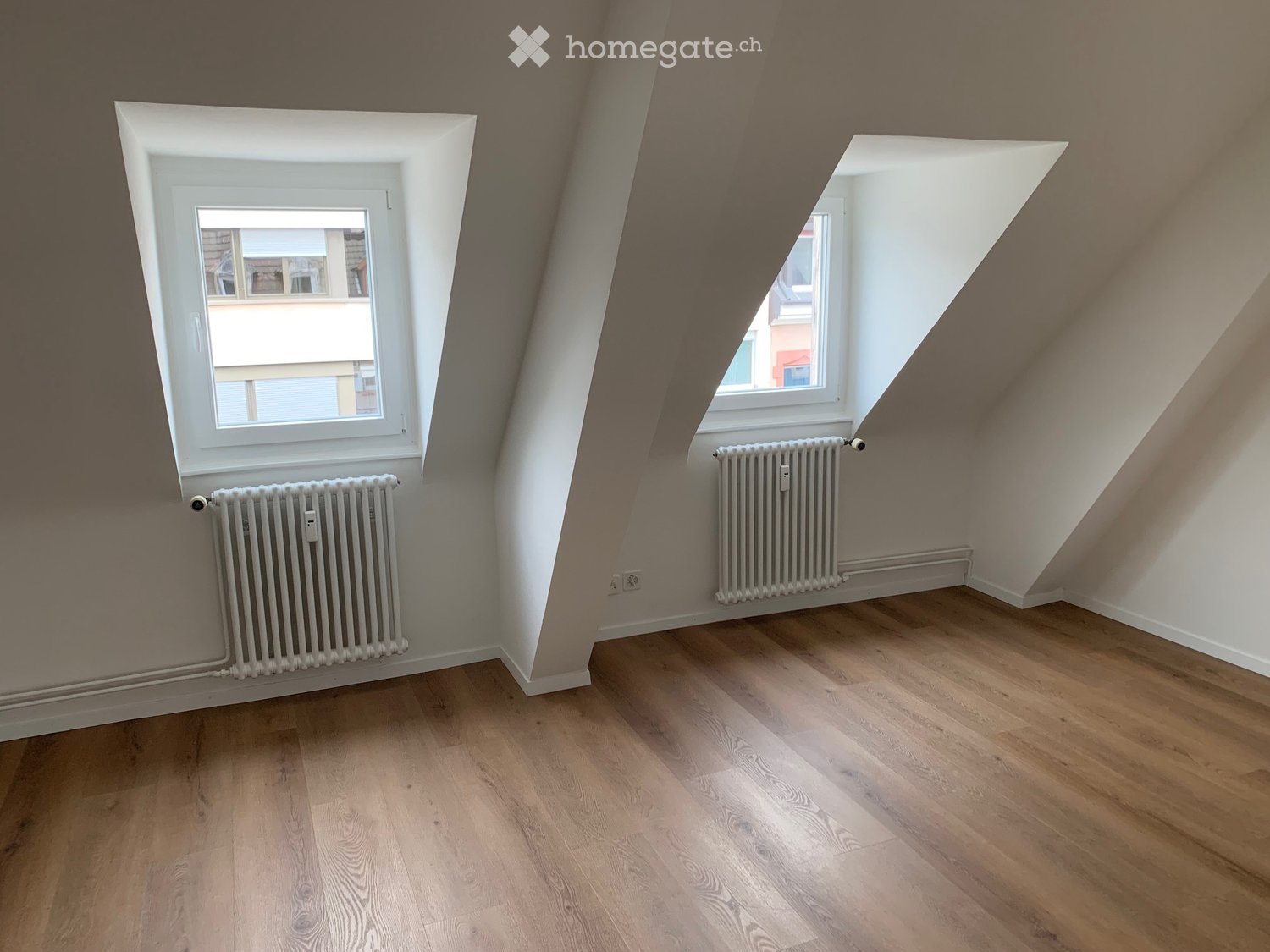 Empty attic room with wooden flooring, white walls, and two windows with radiators below them.