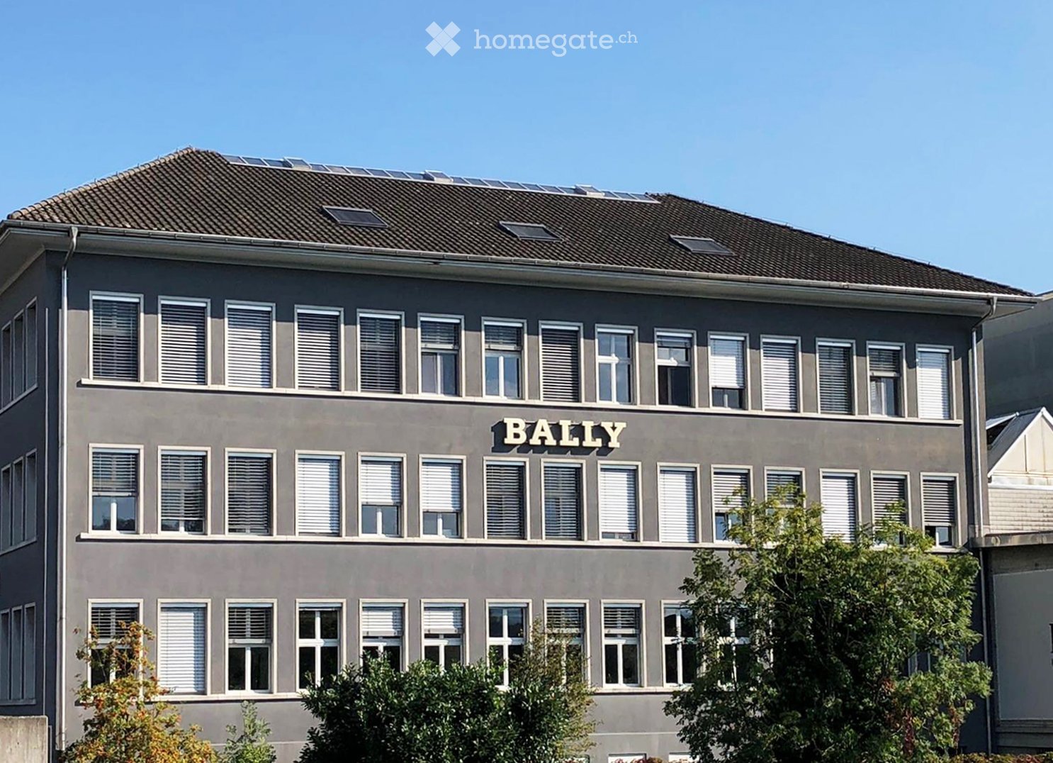 Three-story building with grey facade, multiple windows with white blinds, and a sign reading "BALLY" on the upper floor.
