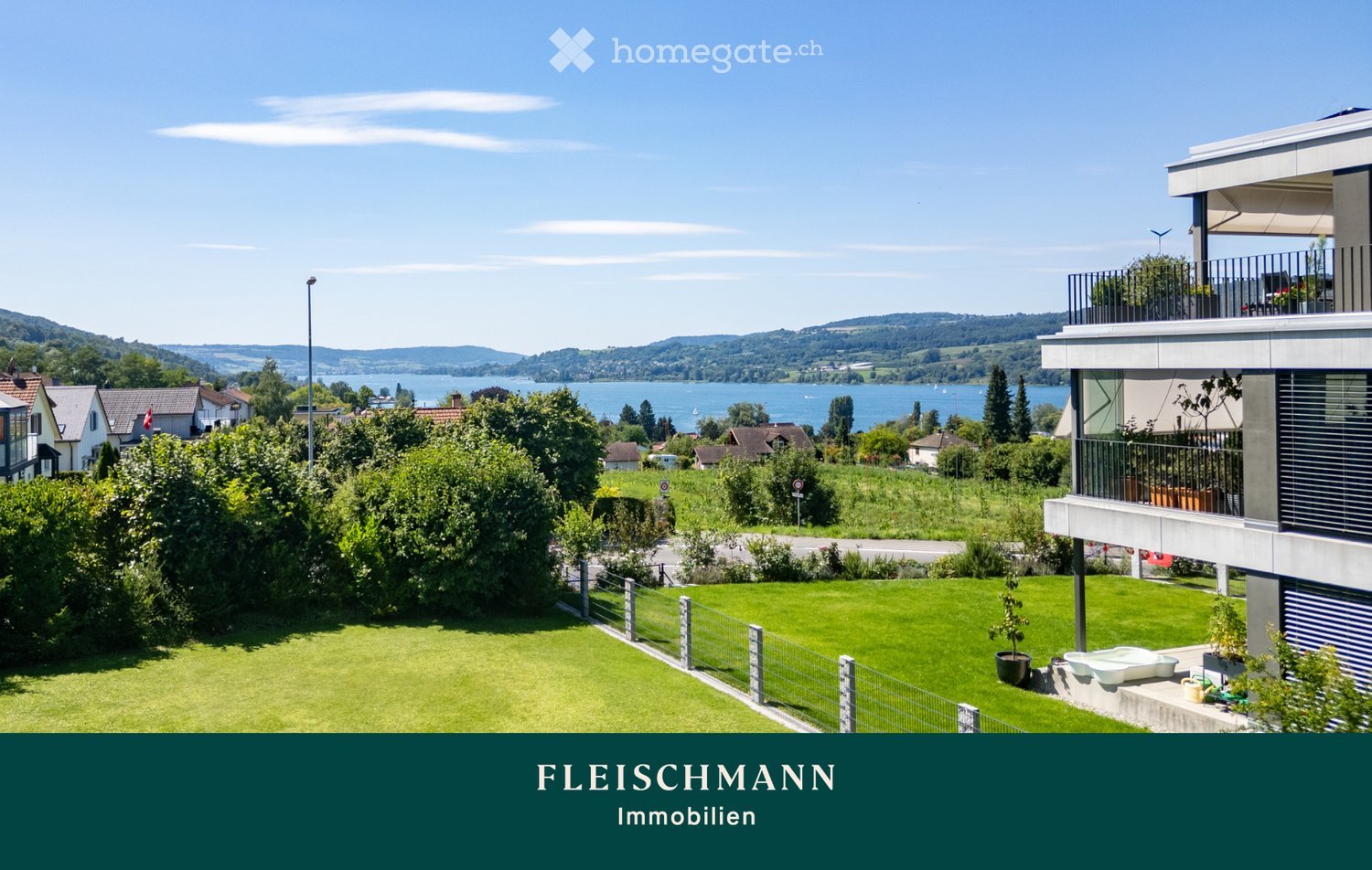 A well-maintained garden with a green lawn, a modern house with a balcony, and a scenic view of a lake and surrounding hills.