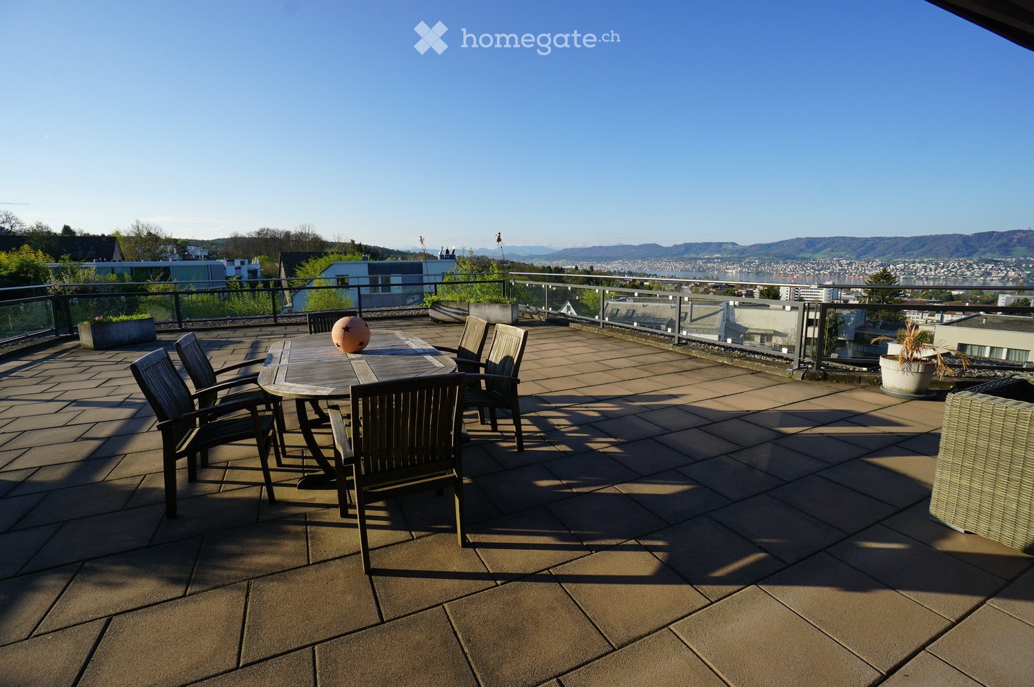 Outdoor dining table with chairs, potted plants, tiled floor, metal railing, distant city view