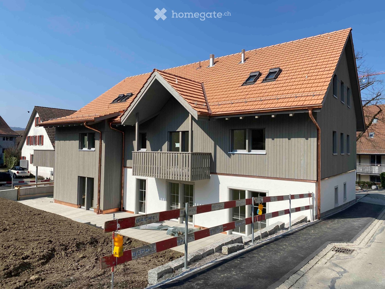 Two-story house with a red-tiled roof, grey siding, and multiple windows, including a balcony on the upper level. The house is partially under construction with barriers and dirt visible in the foreground.