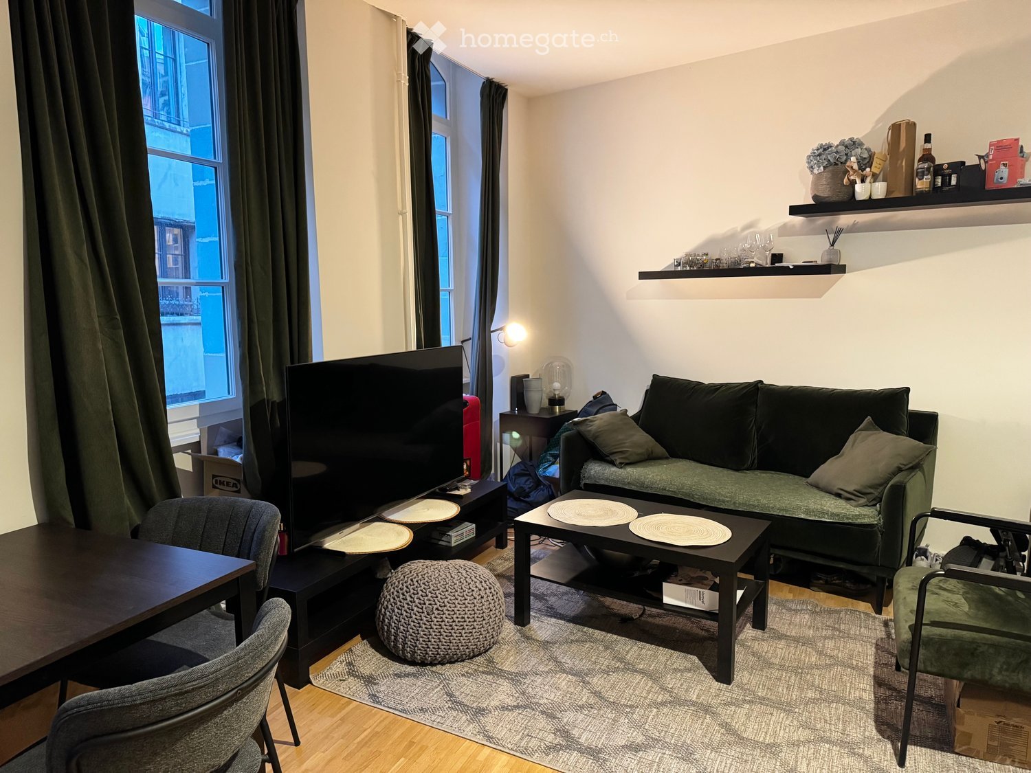 A modern living room with a grey sofa, coffee table, round wooden table, and a large television. The room features wooden flooring, large windows with dark curtains, and shelves with decorative items.