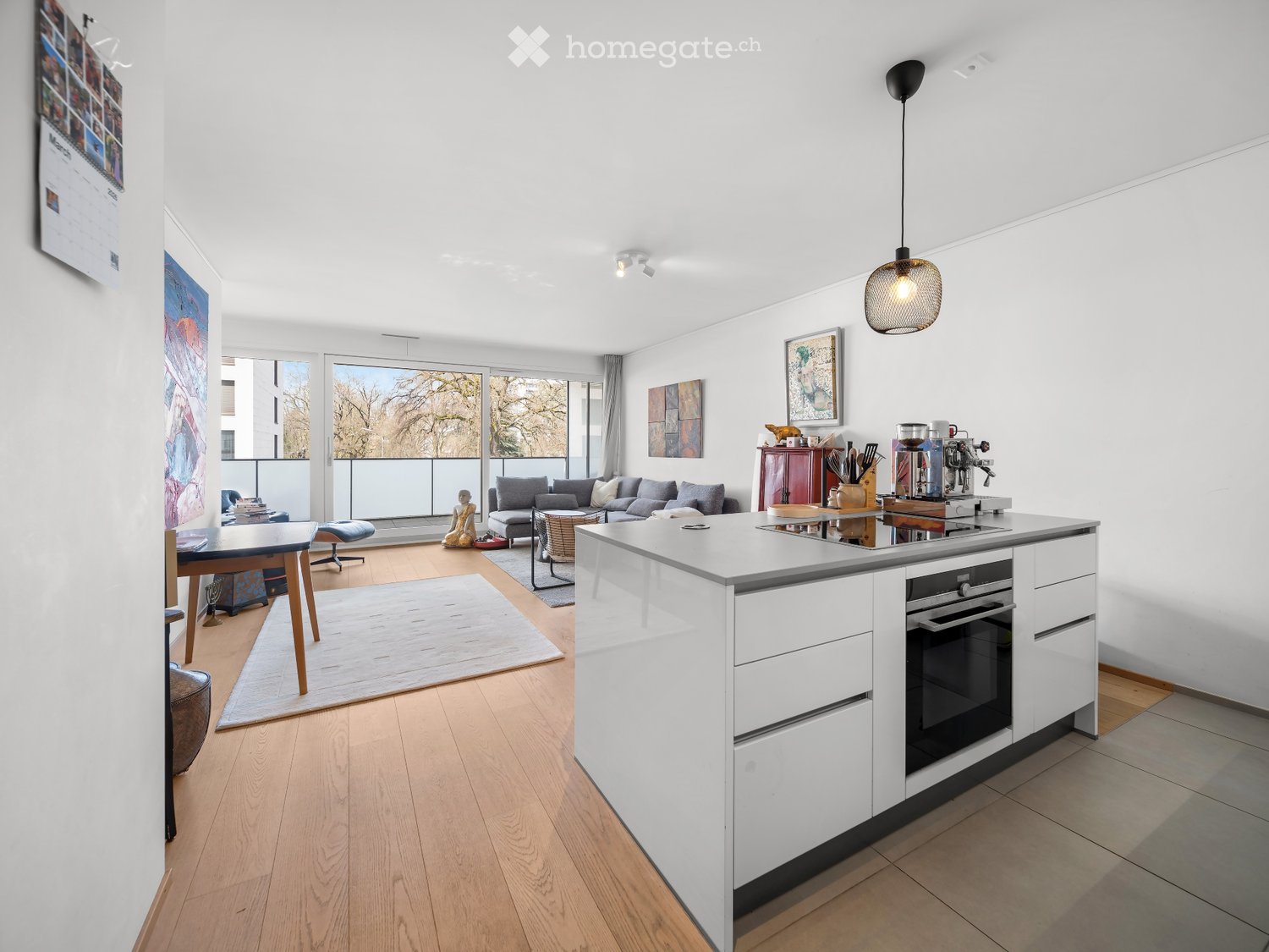 Open-plan living and kitchen area with white walls, wooden flooring, large windows, a sofa, coffee table, kitchen island, and various appliances.
