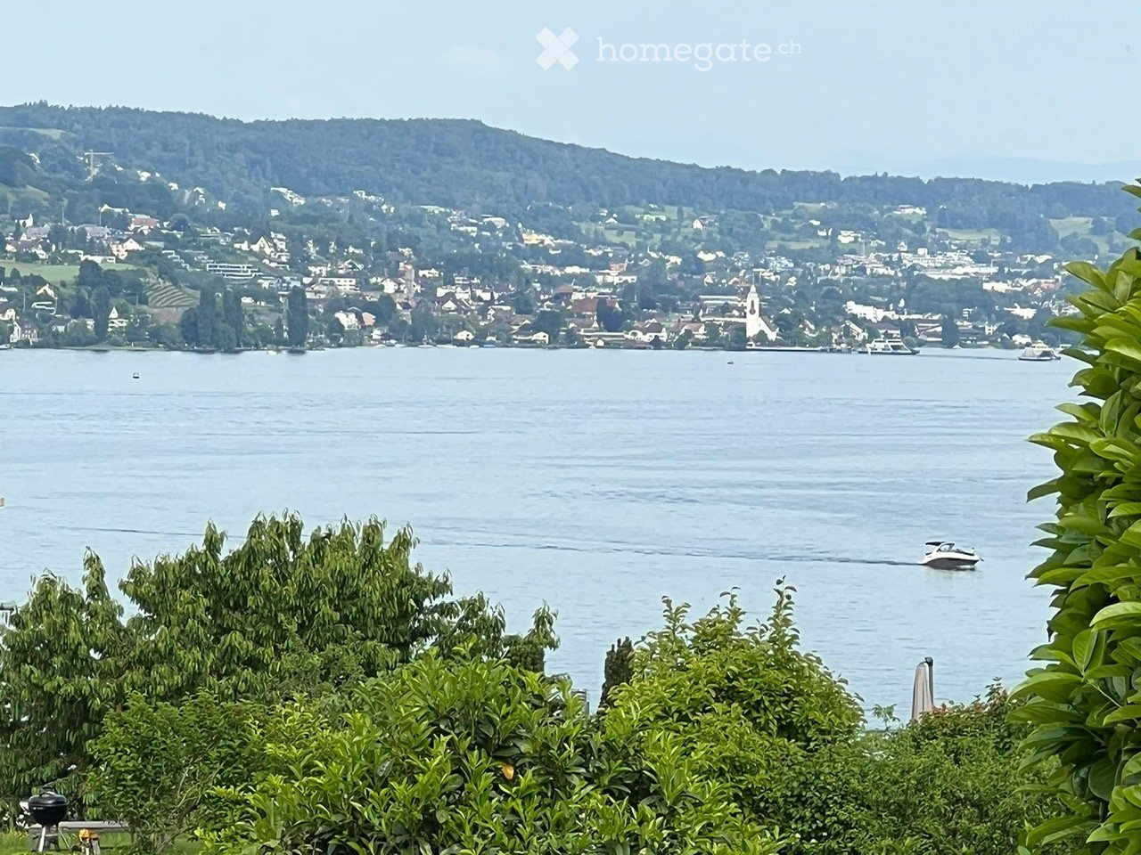 Scenic view of a town across a lake with boats, surrounded by green hills and trees.