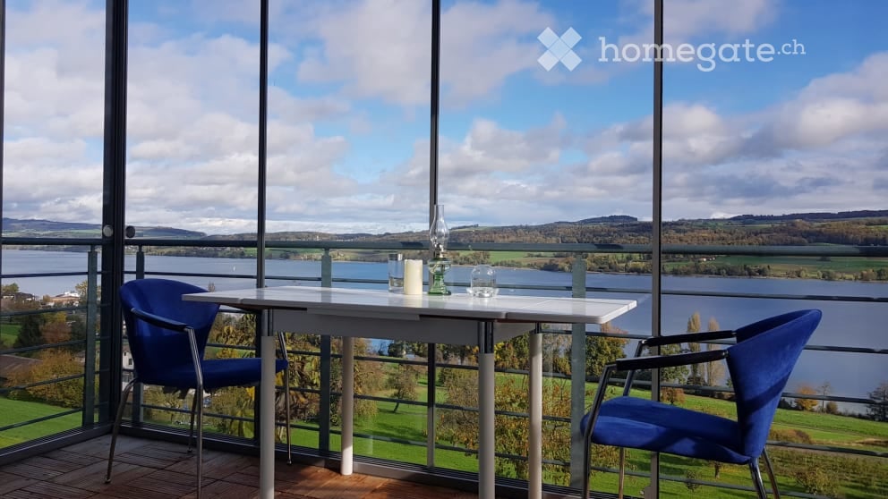 Balcony with a scenic view of a lake and surrounding landscape, featuring a white table, two blue chairs, a candle, and a decorative vase.