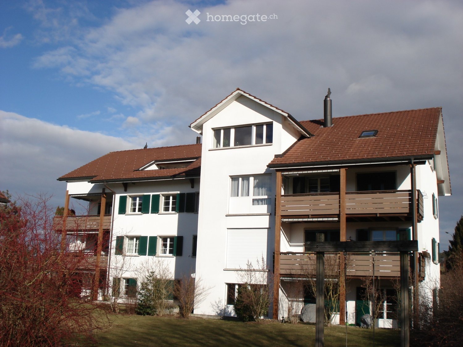 Two-story white house with red-tiled roof, multiple windows with green shutters, balconies, and a chimney.