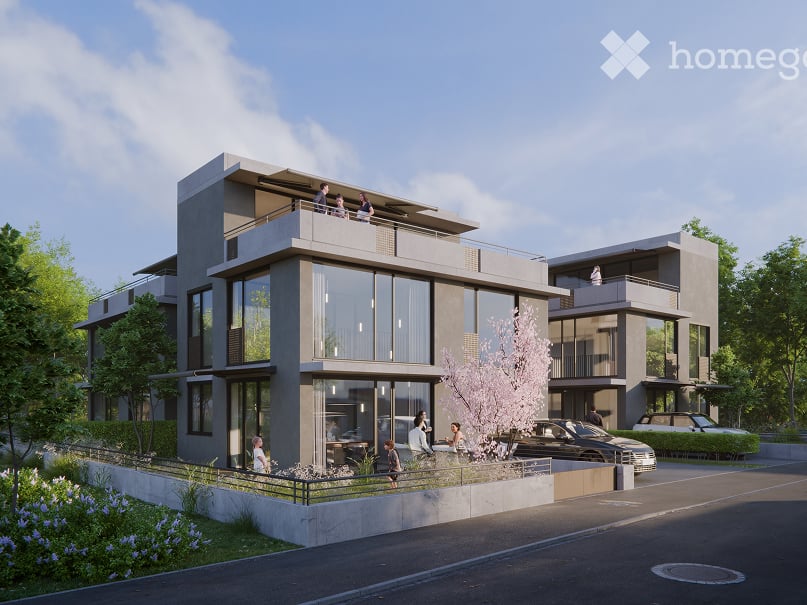 Modern two-story residential building with large glass windows, balconies, and contemporary design. The building is surrounded by greenery and has a parking area in front.